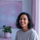 A smiling woman in her late 30s with dark hair, looking directly at the camera. The background is softly blurred with hints o