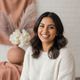 Smiling South Asian woman in her late 20s, facing forward, with a warm, trustworthy expression. Background is blurred with te