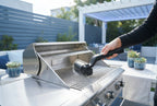 Person using a cordless electric grill brush to clean a stainless steel barbecue on a modern patio.